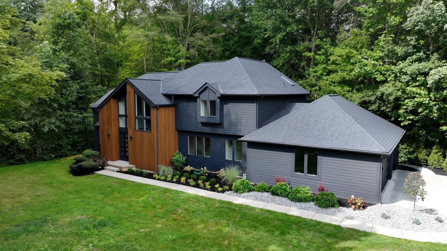 Aerial view of a modern two-story home with wood and charcoal siding, new landscaping, and a wooded backdrop